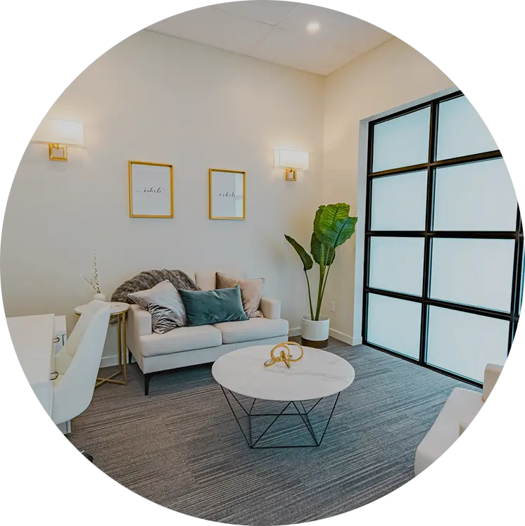 A cozy room with a loveseat, a plant in the corner, a round table in the middle, and frosted windows